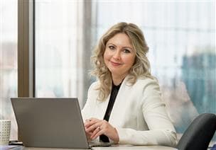 Photo portrait d’une employée souriant à la caméra dans la salle de conférence de l’ÉLHD au Suncor Energy Centre à Calgary, en Alberta. Elle a un ordinateur portable ouvert et on perçoit des gratte-ciels flous par la fenêtre derrière elle.