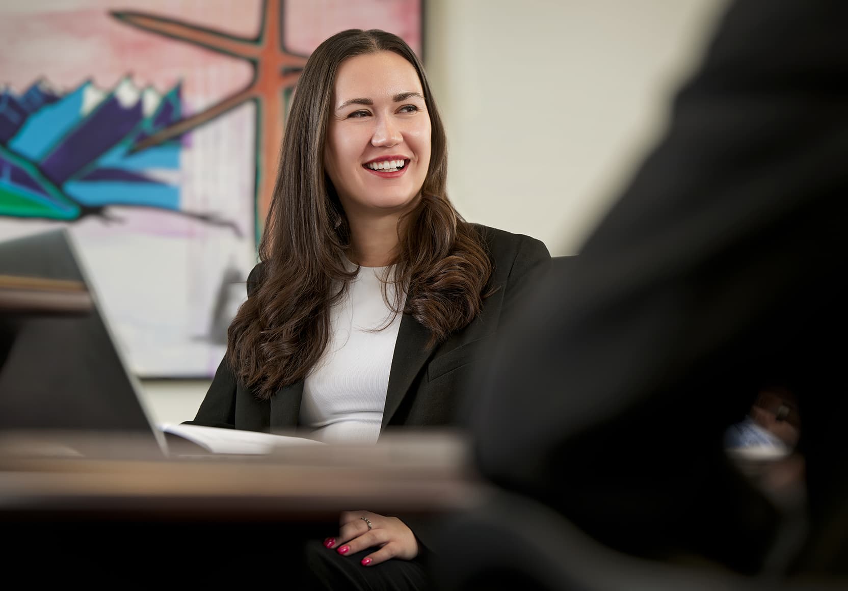 A female employee sitting at a table in a meeting in the ELT Boardroom at the Suncor Energy Centre. Another employee's elbow is blurred out in the foreground