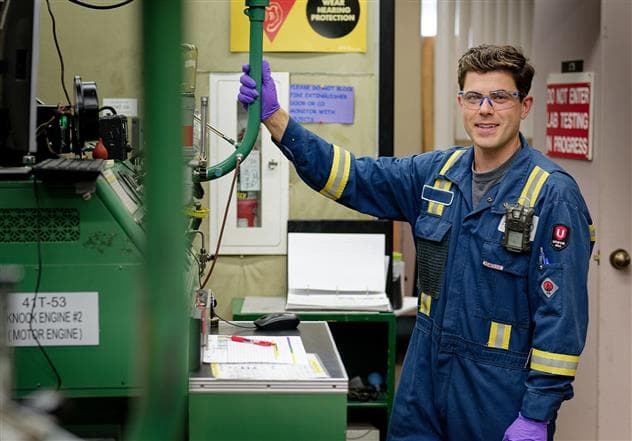A male lab technician working in the Edmonton Refinery lab beside a green motor engine. He is wearing safety glasses, coveralls and gloves.