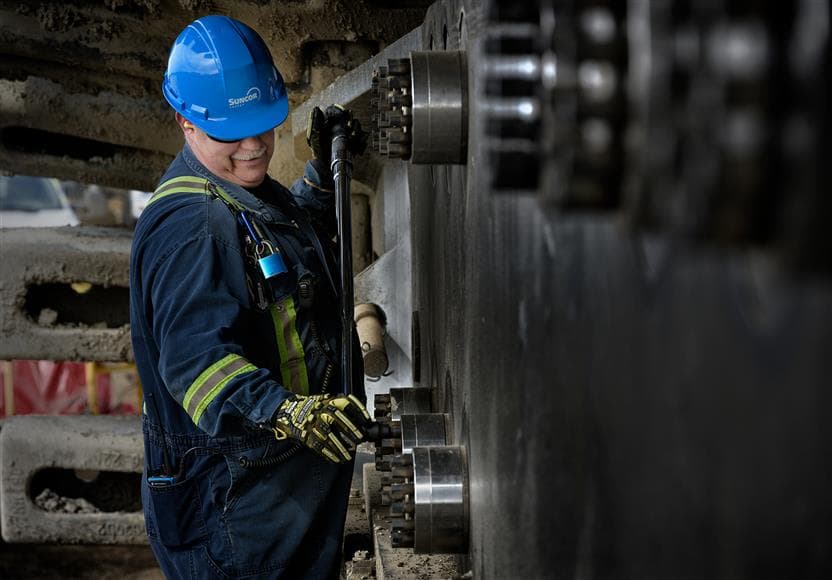 A male worker is working on an electric shovel track on the Borealis Shovel Pad at Base Plant. He is wearing a blue hard hat, ear plugs, coveralls and safety glasses.