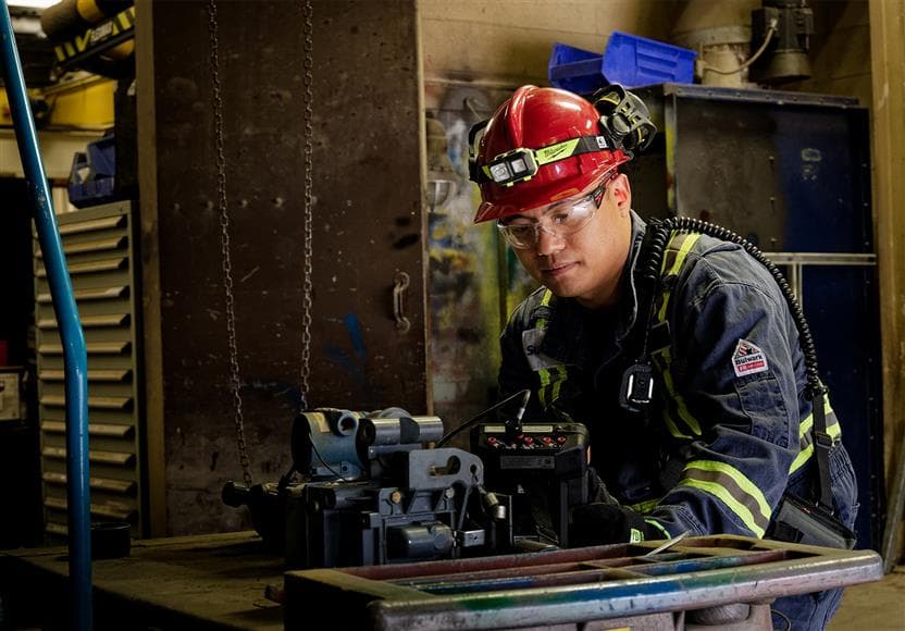 A Suncor employee working in the Edmonton Refinery Machine shop. He is wearing a red hard hat, safety glasses, coveralls and gloves.
