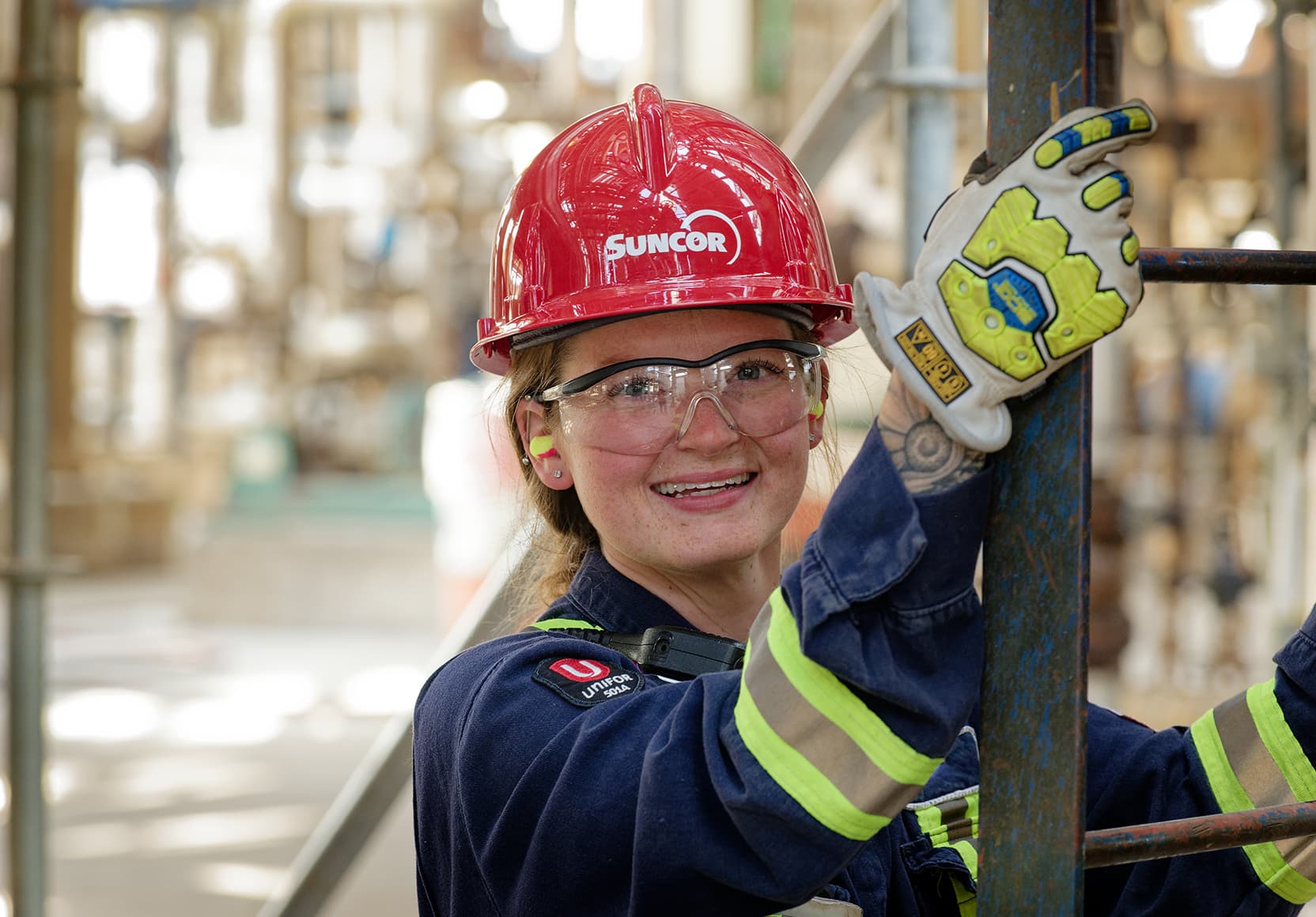 A headshot of a young employee standing with her hands on a ladder at the Edmonton Refinery. She is wearing a red hard hat, safety glasses, ear plugs, coveralls and gloves.