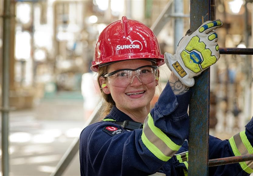 Photo portrait d'une jeune employée dont les mains sont posées sur une échelle à la raffinerie d'Edmonton. Elle porte un casque rouge, des lunettes de sécurité, des bouchons d'oreille, une combinaison et des gants.
