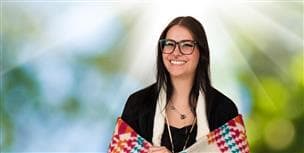 A headshot of a female Indigenous student. She is wrapped in a traditional blanket and is holding a feather. There is a vibrant array of colors in the background. 