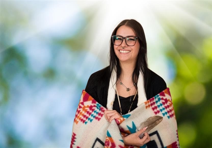 Photo portrait d’une étudiante autochtone. Elle est enveloppée dans une couverture traditionnelle et tient une plume. Il y a une gamme de couleurs vibrantes en arrière-plan. 