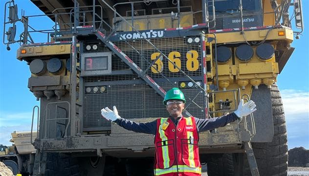 A co-op student standing in front of a Komatsu truck at the Base Plant site near For McMurray. He is wearing PPE and is smiling with his arms in the air.