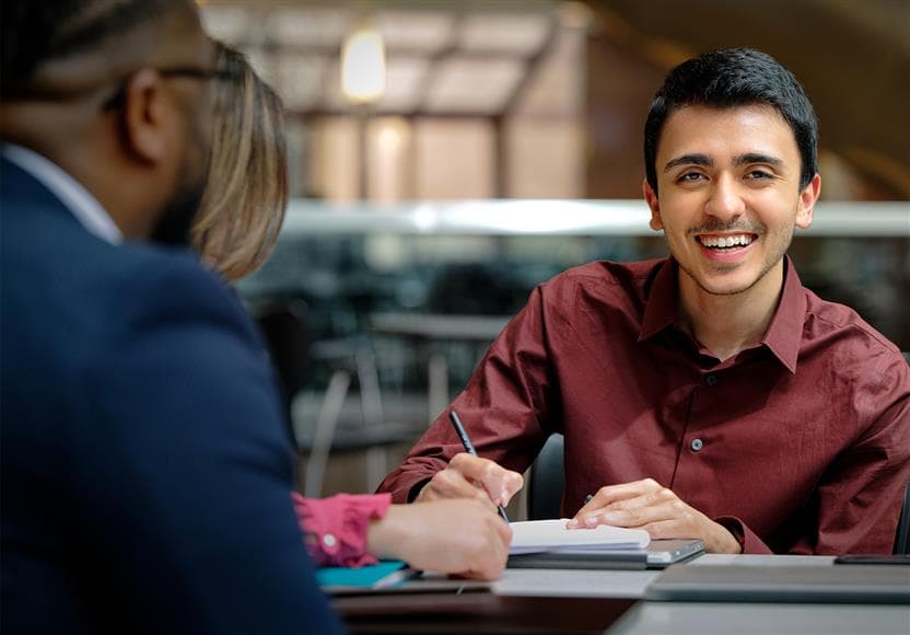 An employee writing in a notebook and smiling at the camera. They are sitting at a table with two other employees in the Suncor Energy Centre lobby.