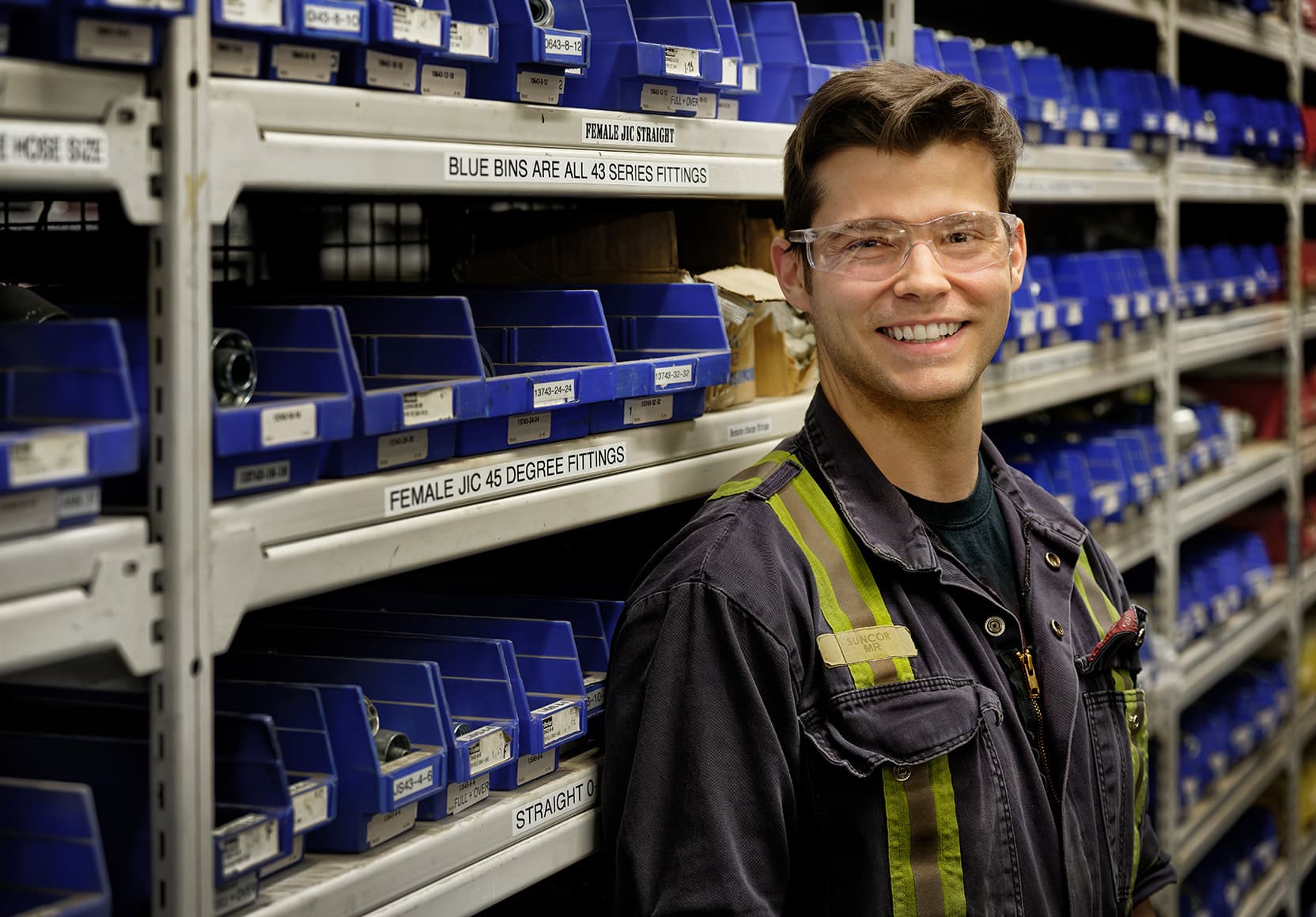 A worker standing in front of shelving with blue bins on it in the truck shop warehouse at Base Plant. He is wearing safety glasses and coveralls.