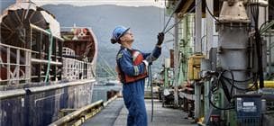 Un travailleur au quai du terminal Burrard. Il tient deux cordes dans ses mains et porte un casque de sécurité bleu, des lunettes de sécurité, des gants, un gilet de sauvetage et une combinaison.