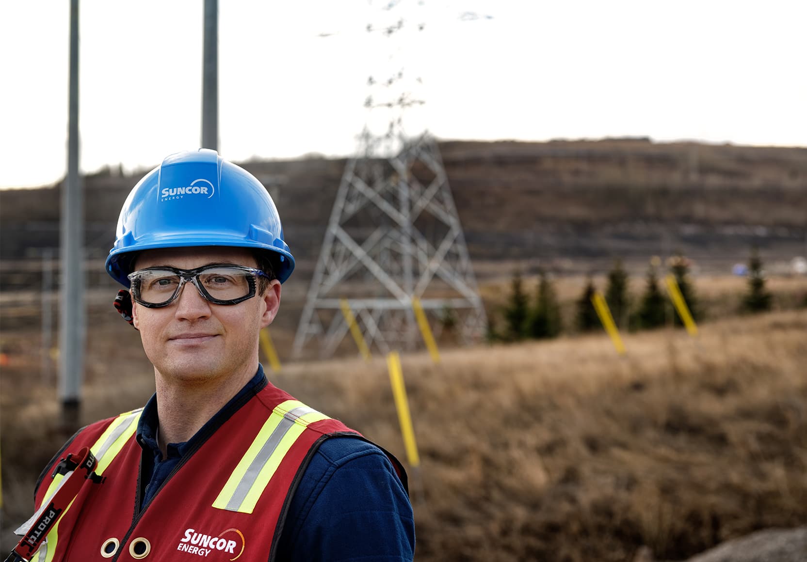 A headshot of a worker outside at Base Plant. He is wearing a blue hard hat, safety glasses and a high-visibility vest. There is a transmission tower out of focus in the background.