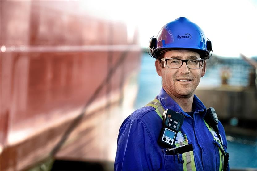 A Suncor employee standing on the dock at the Sarnia Refinery. He is wearing a blue hard hat, safety glasses and coveralls. The river is blurred out in the background.