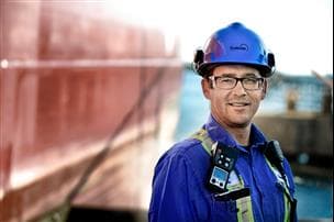 A Suncor employee standing on the dock at the Sarnia Refinery. He is wearing a blue hard hat, safety glasses and coveralls. The river is blurred out in the background.