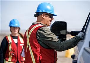 Deux travailleurs à l’extérieur au point d’entrée D de la mine de l’usine de base. L’employé a une main posée sur la fenêtre d’un camion blanc et parle au conducteur. Ils portent des lunettes de sécurité, un casque de sécurité bleu, des gants et une veste de sécurité.