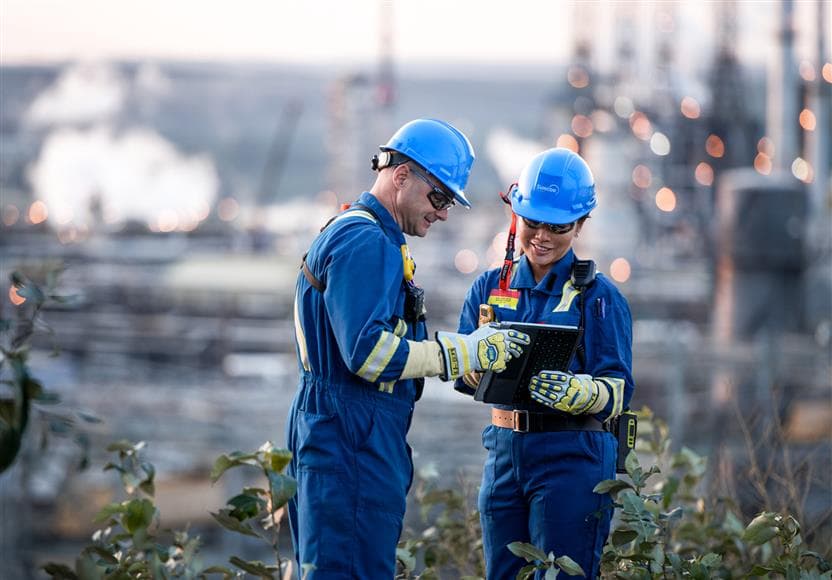 Two workers outside in PPE looking at a tablet together. Base Plant is in the background. 