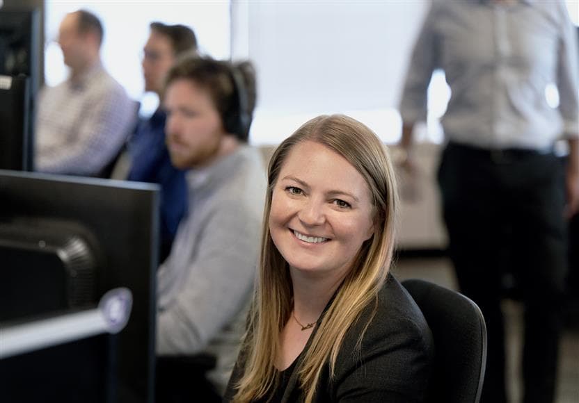 Une employée travaillant au Suncor Energy Centre. Elle sourit à la caméra et est assise à un bureau, et il y a trois employés dont l’image est floutée en arrière-plan.