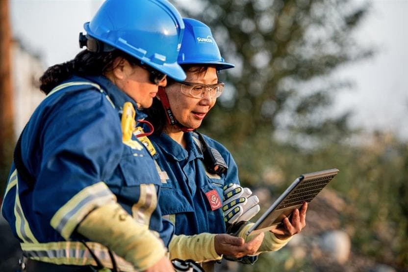 Two Suncor workers look at a tablet together. They are wearing hard hats, safety glasses, and coveralls, and trees can be seen in the background. 