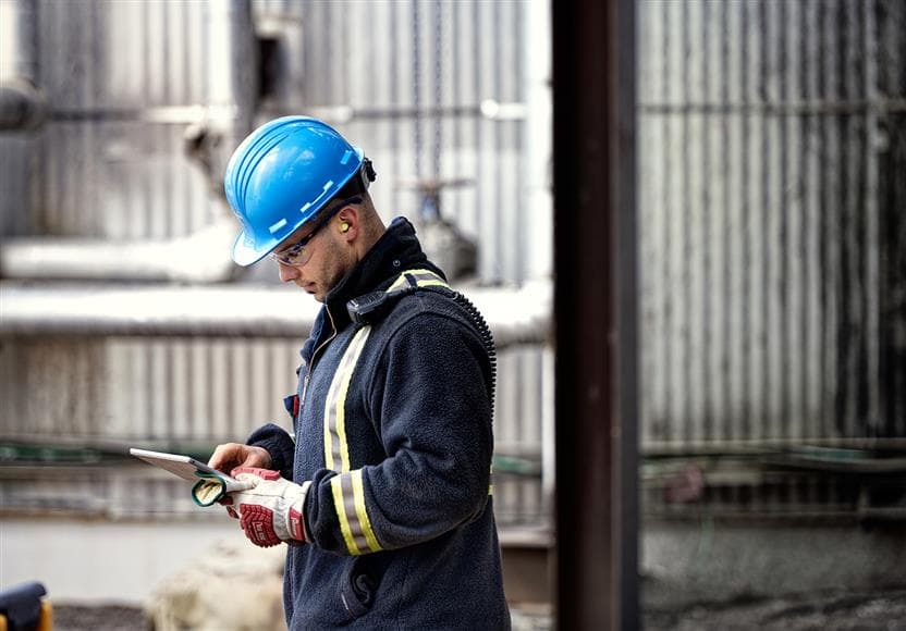 A Suncor worker using a Microsoft Surface Pro at MacKay River Plant 300. He is wearing a blue hard hat, safety glasses, ear plugs, coveralls and gloves.
