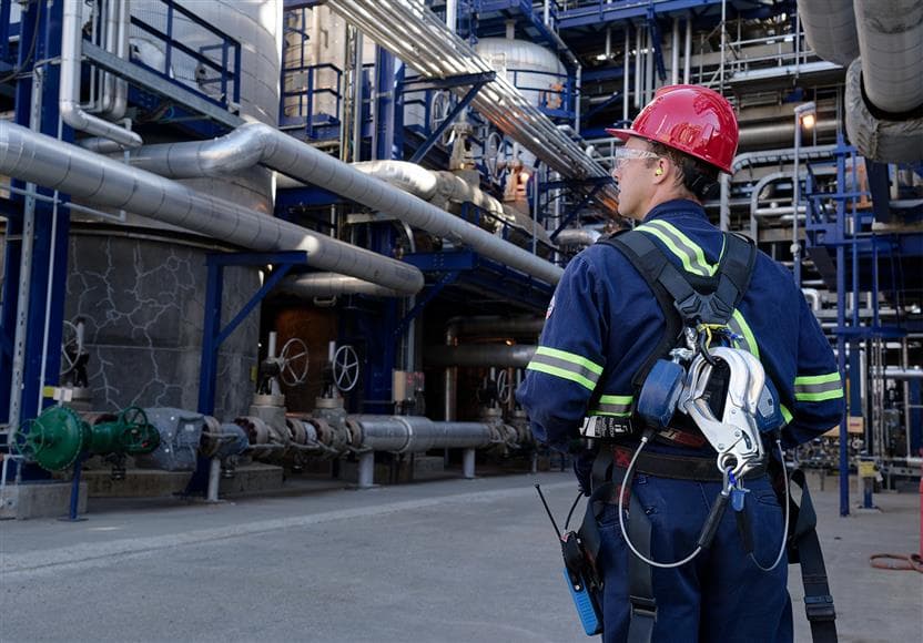 A male worker standing and facing away from the camera. He is wearing a red hard hat, safety glasses, ear plugs, and coveralls.
