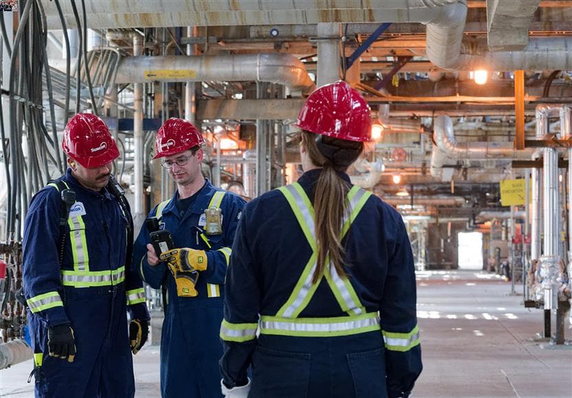 Trois travailleurs se tenant à l’usine 10 à la raffinerie d'Edmonton. Ils portent un casque de protection rouge, des lunettes de sécurité, des bouchons d'oreilles, une combinaison et des gants. Deux d’entre eux regardent un appareil, tandis que l’autre travailleuse a le dos tourné à la caméra.