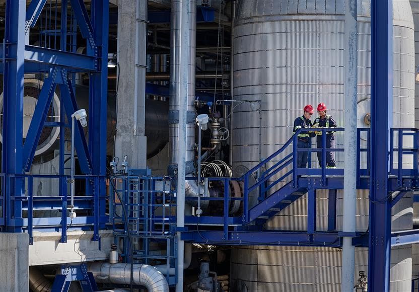 Two workers standing on a walkway by some stairs at the Edmonton Refinery sulphur plant. They are wearing red hard hats, safety glasses, ear plugs, coveralls and gloves. They are looking at a tablet that one of them is holding.