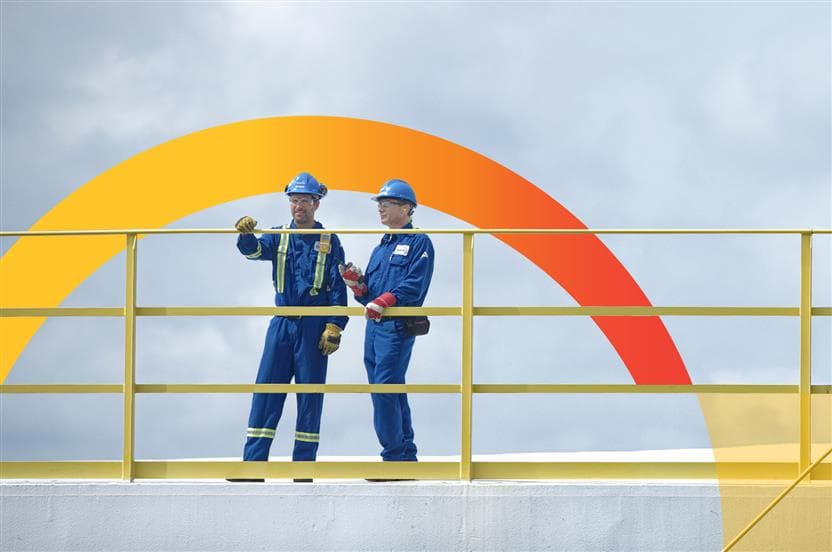 Two male workers standing on top of a storage tank at the Montreal refinery. They are wearing blue hard hats, safety glasses, coveralls and gloves.