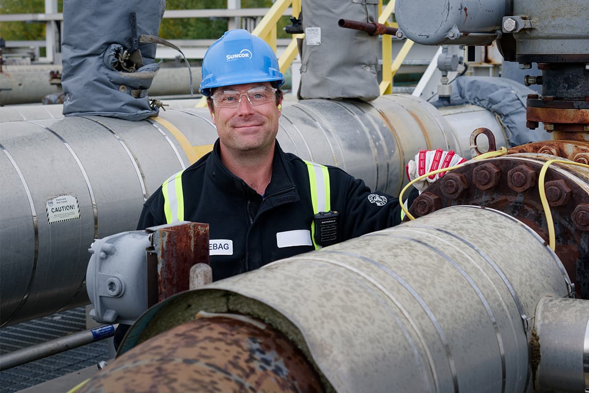 Suncor employee at Firebag Terminus One. He is smiling at the camera and wearing a blue hard hat, safety glasses, gloves and ear plugs.