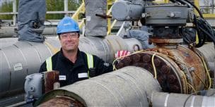 Suncor employee at Firebag Terminus One. He is smiling at the camera and wearing a blue hard hat, safety glasses, gloves and ear plugs.