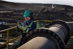 A Suncor worker using a site phone outside by a pipeline at Suncor's Base Plant in Northern Alberta. She is wearing a green hard hat, ear plugs, coveralls, gas detector and safety glasses. There are an electric shovel and haul truck out of focus in the background.