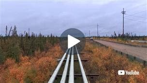 At Suncor’s Firebag site, a wide outdoor view of three metal pipelines running straight through autumn vegetation toward the horizon. A gravel road with power poles runs alongside on the right. A large play button appears in the center of the image, with a YouTube logo in the bottom right corner.