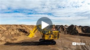 The first PC9000, Komatsu's largest hydraulic shovel commercially available in the world, shown at Suncor’s Fort Hills mine. The shovel is decorated with a Canadian flag and a Suncor flag, and cloudy blue skies paint the background. A large play button appears in the center of the image, with a YouTube logo in the bottom right corner.