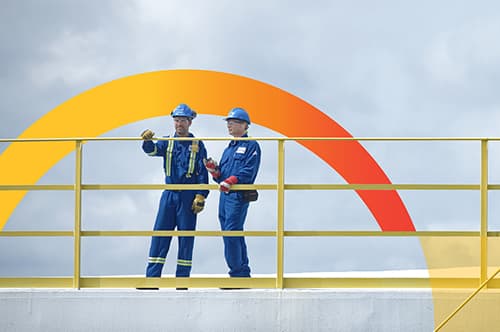 Two male workers standing on top of a storage tank at the Montreal refinery. They are wearing blue hard hats, safety glasses, coveralls and gloves.