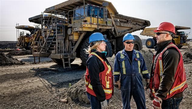 Une travailleuse et deux travailleurs se tenant à l'extérieur à l'usine de base. Ils portent de l'ÉPI et discutent entre eux. Il y a quatre camions de transport en arrière-plan.