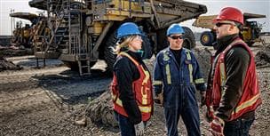 Une travailleuse et deux travailleurs se tenant à l'extérieur à l'usine de base. Ils portent de l'ÉPI et discutent entre eux. Il y a quatre camions de transport en arrière-plan.
