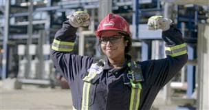 Une employée de Suncor à l’usine de base sourit et lève les bras pour célébrer. Elle porte une combinaison, des lunettes de protection et un casque de protection rouge arborant le logo de Suncor. On peut apercevoir de la machinerie en arrière-plan.
