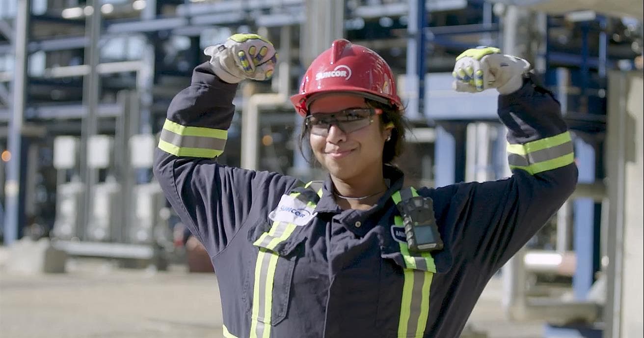 Une employée de Suncor à l’usine de base sourit et lève les bras pour célébrer. Elle porte une combinaison, des lunettes de protection et un casque de protection rouge arborant le logo de Suncor. On peut apercevoir de la machinerie en arrière-plan.