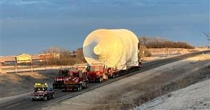 Un énorme objet cylindrique blanc (tambour à coke) est transporté sur deux remorques rouges sur une route à deux voies.  Il y a deux autres remorques et un camion rouges qui suivent le chargement.