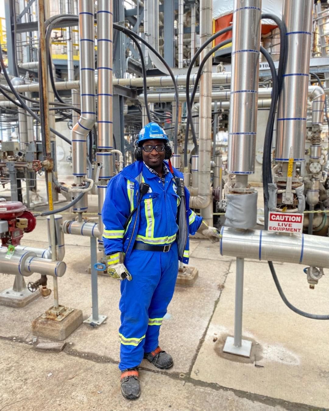 A man wearing blue coveralls, ear protection, large gloves, boots and a hardhat. He is standing near many steel pipes. 