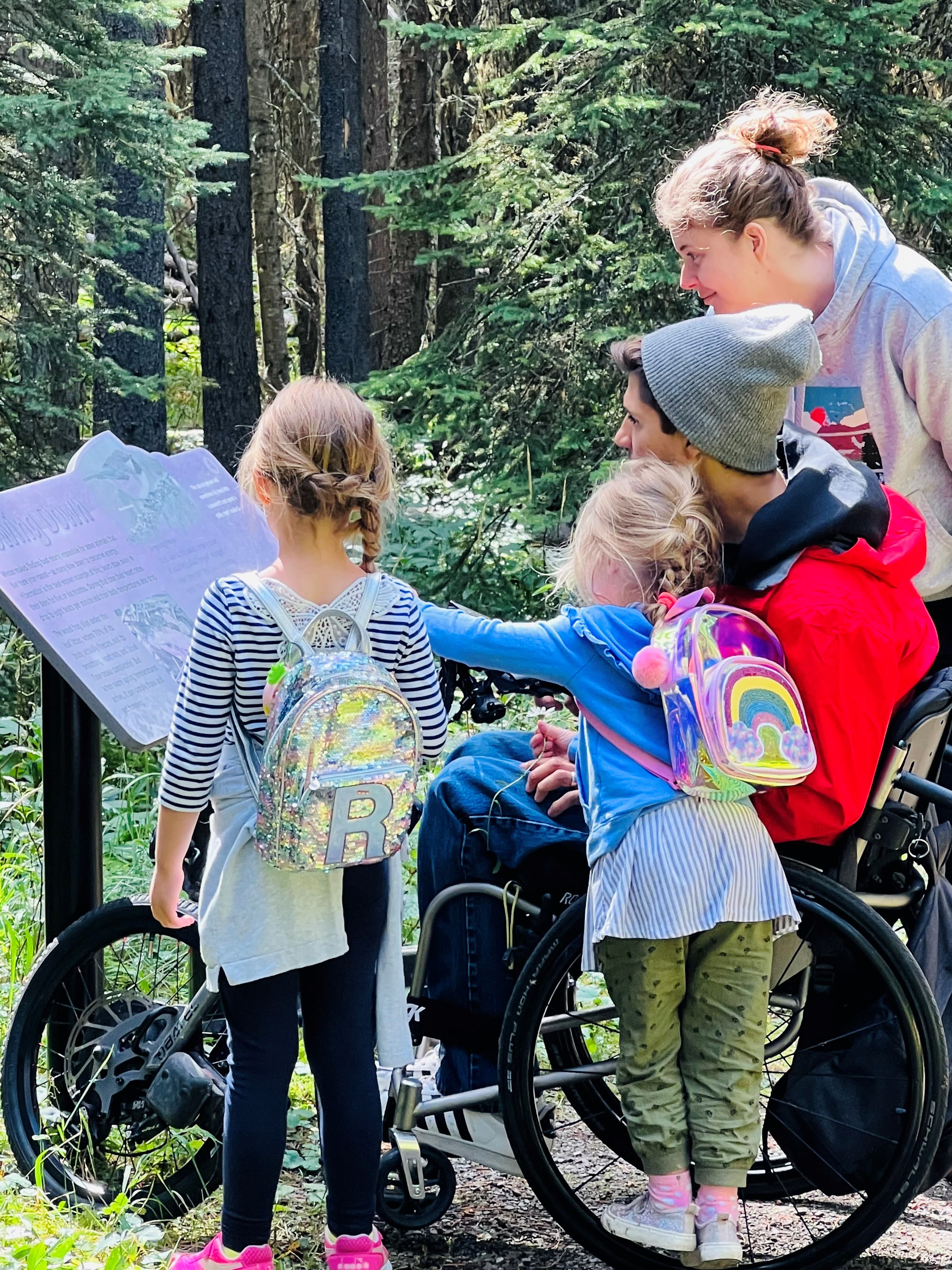 Mike en fauteuil roulant, sa femme et leurs deux filles regardent un panneau sur un sentier de randonnée.
