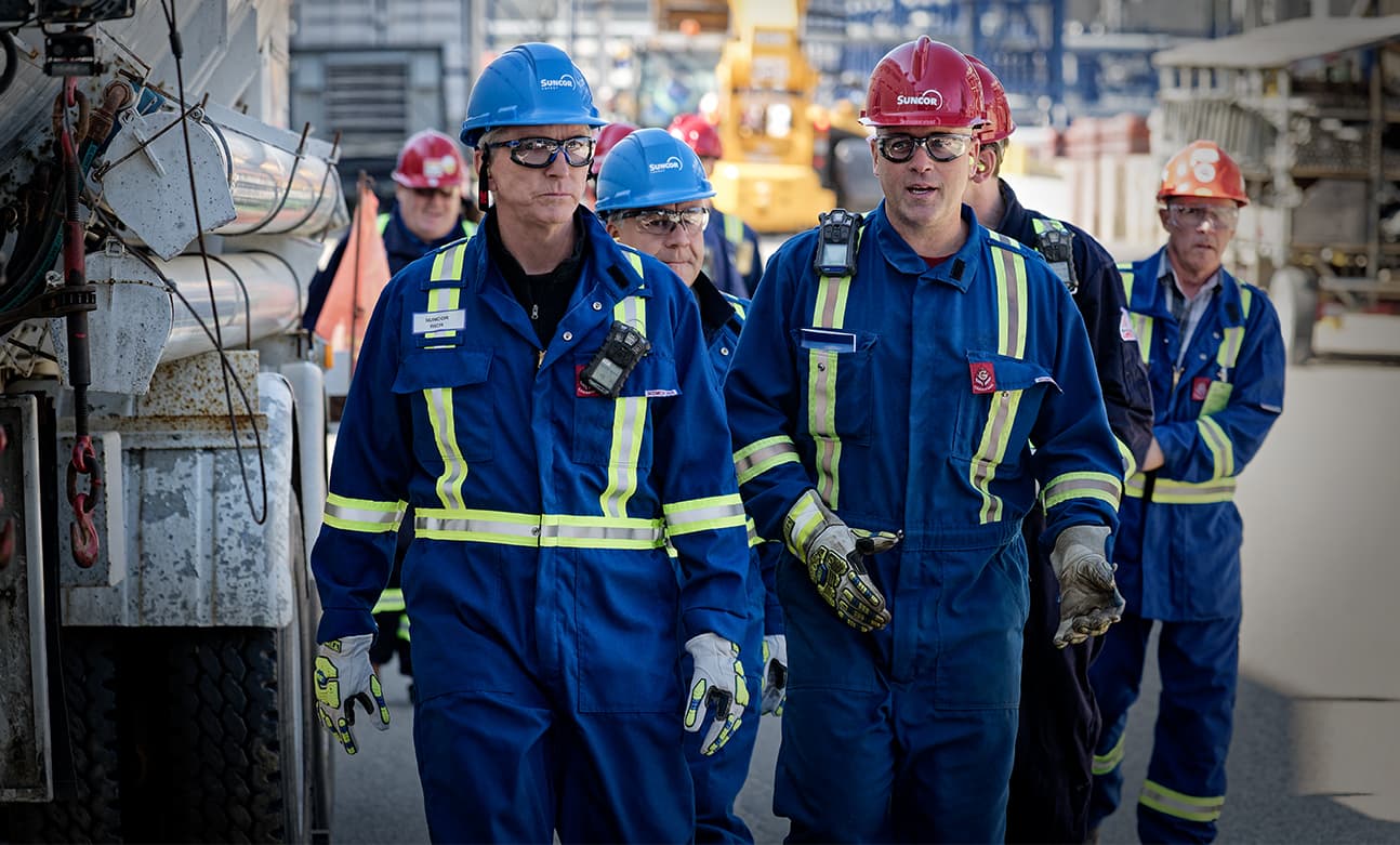 Suncor executives and employees talk while touring the Edmonton Refinery. They are wearing red hard hats, safety glasses, ear plugs, coveralls and gloves.