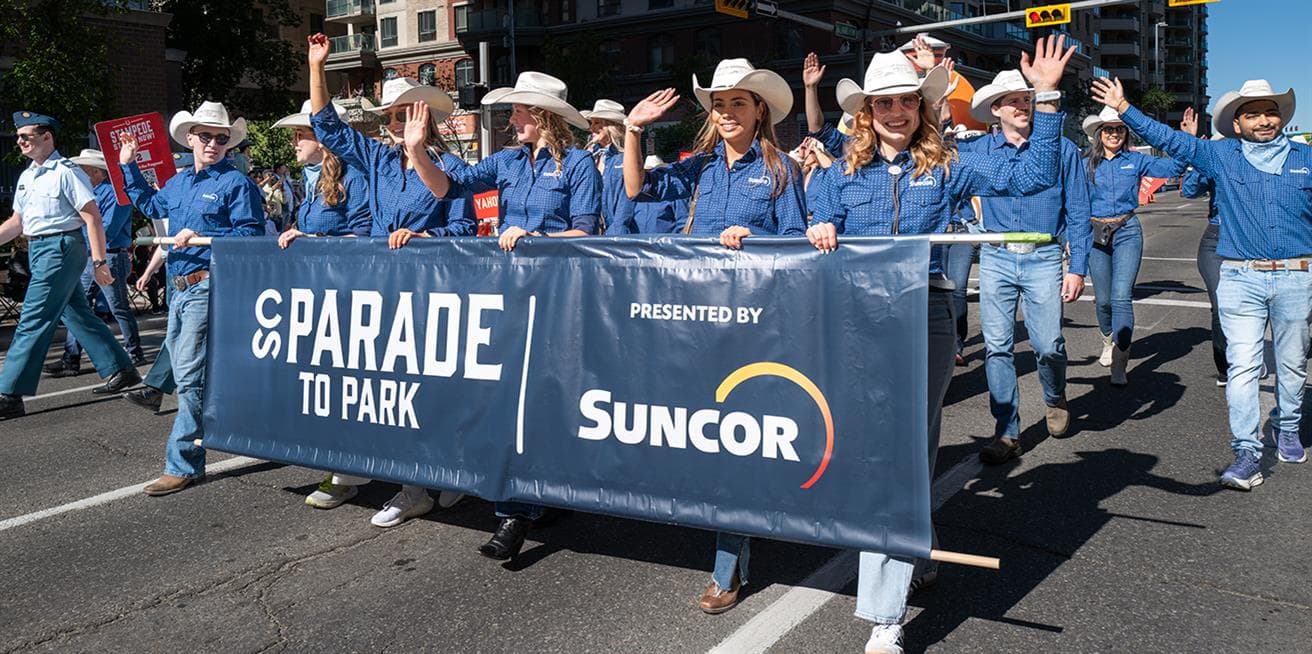 Suncor employees in blue shirts and white cowboy hats carry a "Parade to Park Presented by Suncor" banner while walking in the Calgary Stampede Parade through downtown Calgary.