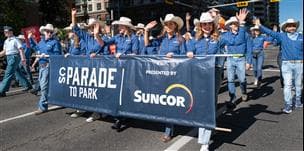 Suncor employees in blue shirts and white cowboy hats carry a "Parade to Park Presented by Suncor" banner while walking in the Calgary Stampede Parade through downtown Calgary.