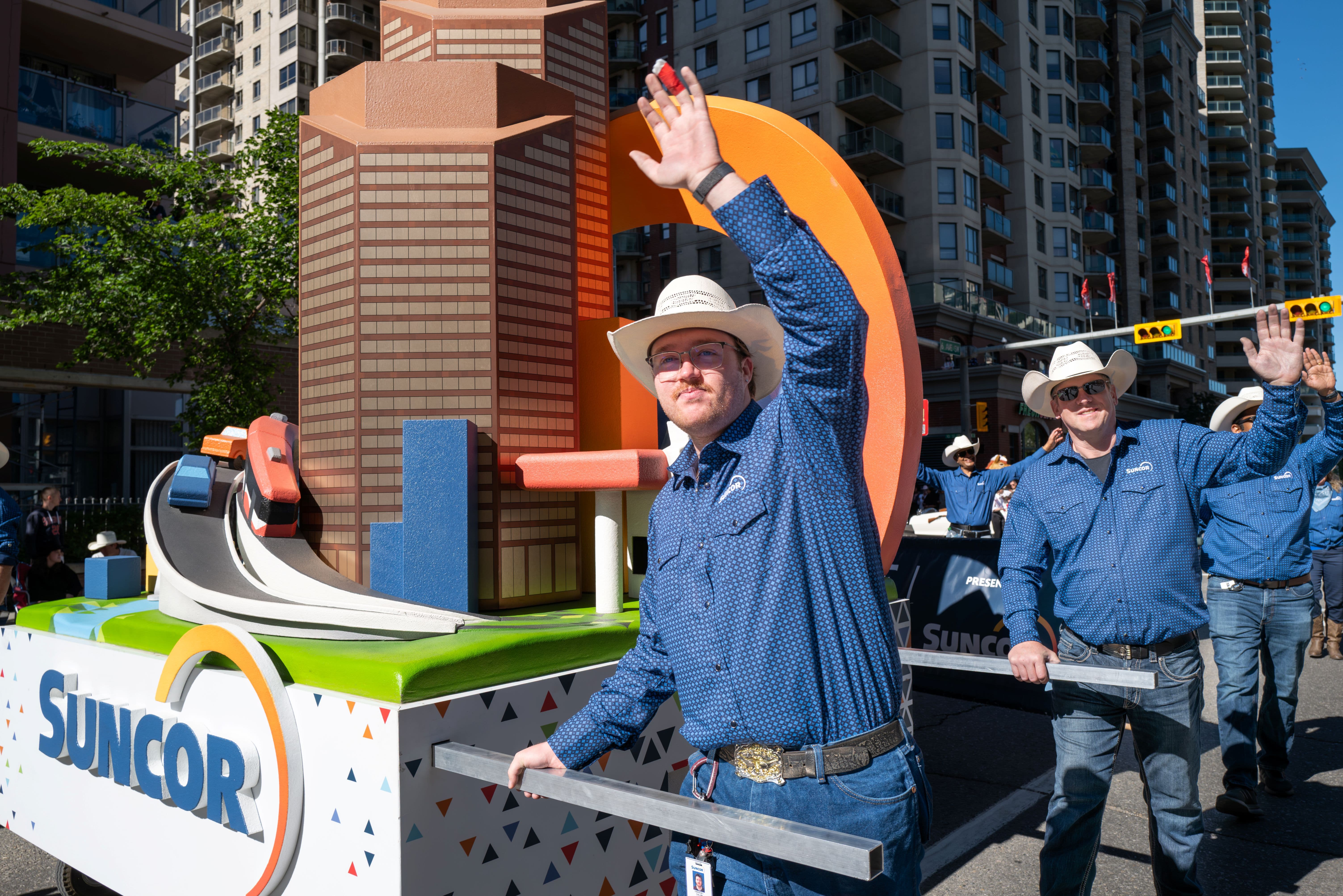 Suncor employee in a blue shirt and white cowboy hat pull a Calgary-themed parade float through downtown Calgary during the Calgary Stampede Parade, waving to spectators along the route