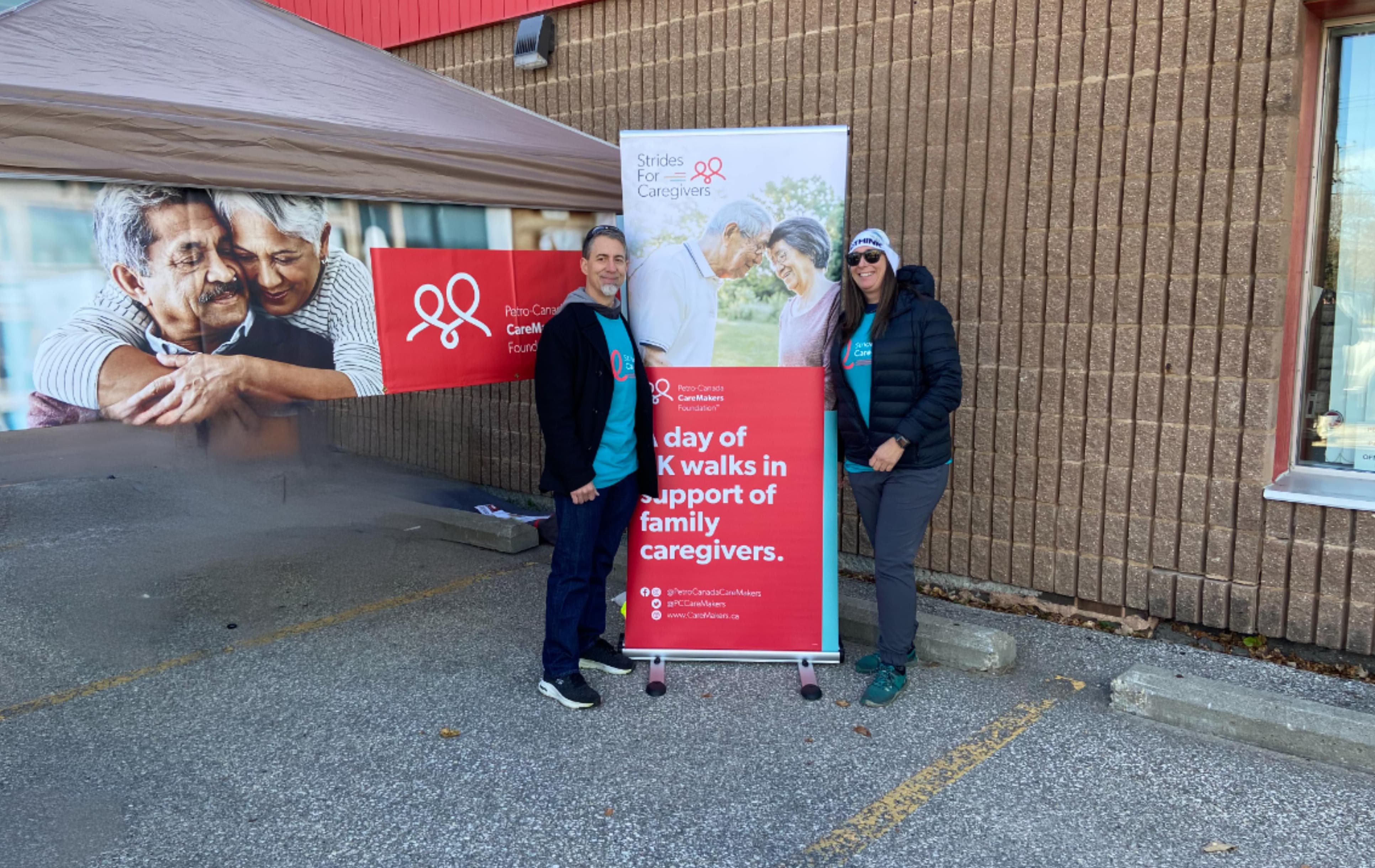 A man and a woman standing beside a Strides for Caregivers banner outside of a brick building.