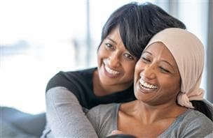 Two women smiling and embracing. One woman is wearing a light coloured scarf on her head.