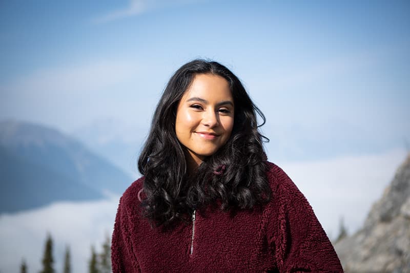 A person with long hair wearing a dark sweater, standing outside with mountains in the background.