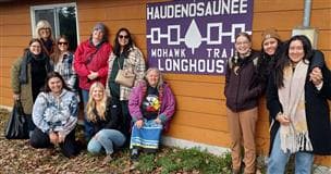 Un groupe de personnes portant des chapeaux, des vestes et des chandails qui se tiennent à l'extérieur d'un bâtiment. Il y a des feuilles sur le sol et une affiche sur le bâtiment indiquant : Haudenosaunee Mohawk Trail Longhouse.