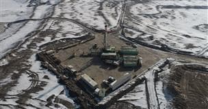 An aerial photo of a drilling rig in a clearing in the woods. The rig is bright green and there are vehicles parked along the perimeter of the clearing. 