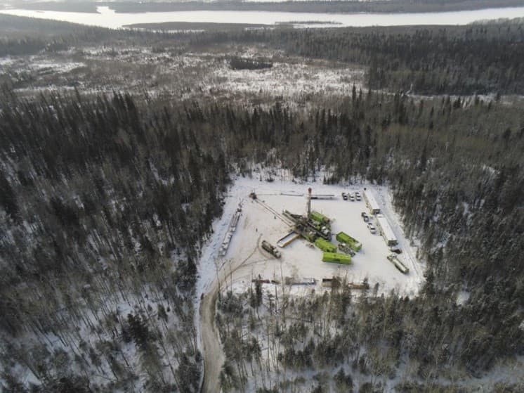 An aerial photo of a clearing where a drilling rig is set up. There is snow on the ground and buildings and vehicles along the clearing perimeter. 