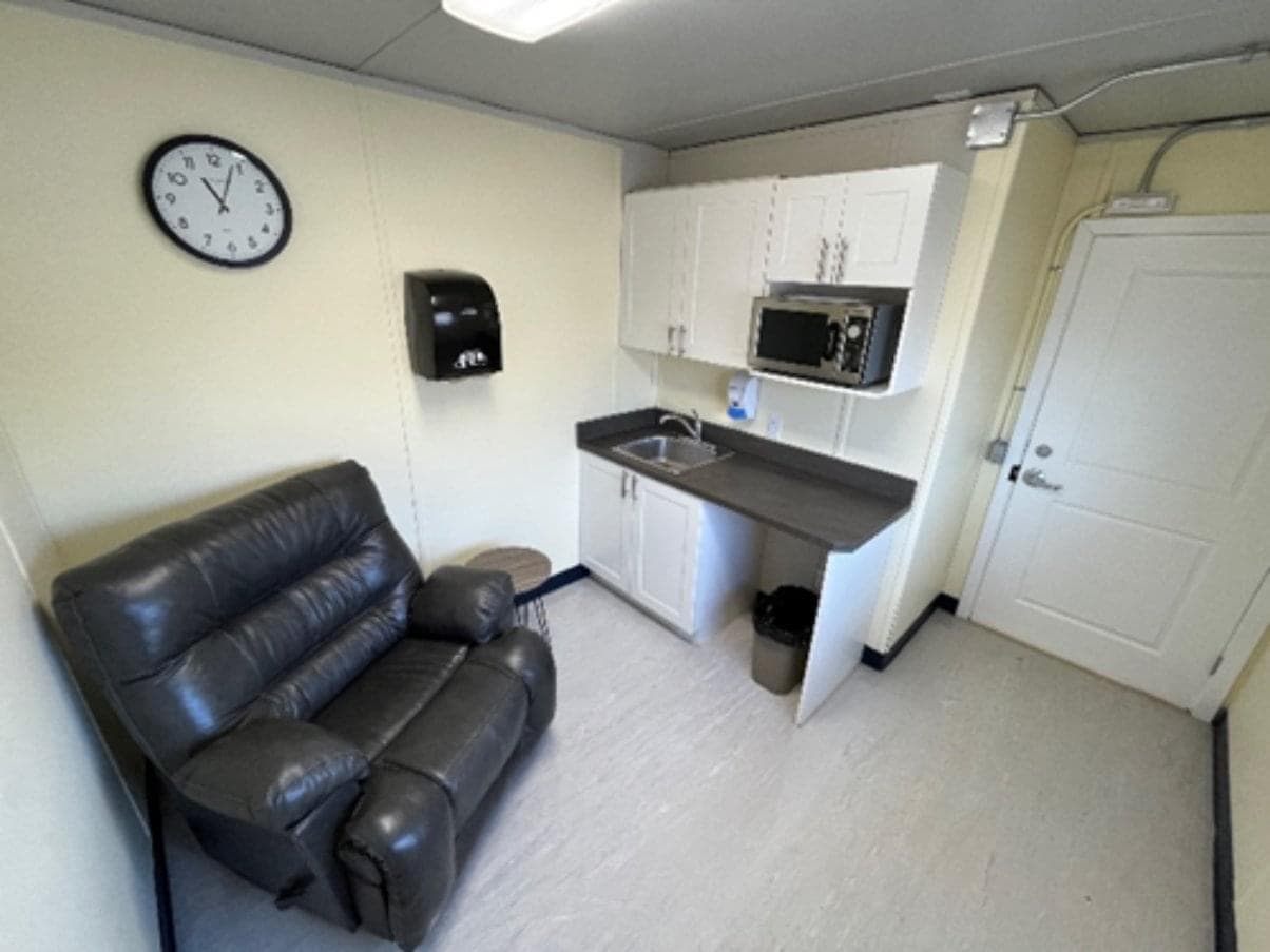 A room with a large reclining chair, a sink, and a microwave. There is a clock on the wall above the chair.