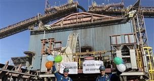 Two men wearing blue coveralls holding balloons stand in front of a plant with large conveyors in the background.